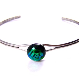 May include: A silver-toned cuff bracelet with a central, round, dichroic glass bead. The bead displays a geometric pattern in shades of green and blue, set against a dark background. The bracelet has a simple, open design.