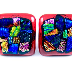 May include: Pair of square-shaped, dichroic glass earrings. The earrings have a deep red base with colorful, iridescent glass fragments in shades of blue, green, gold, and purple. The glass pieces are arranged in a mosaic-like pattern.