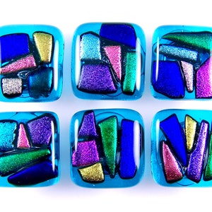 May include: Six square glass tiles with a light blue background. Each tile features a mosaic of colorful glass pieces in shades of blue, purple, pink, green, and gold. The glass pieces have a glittery, textured appearance.