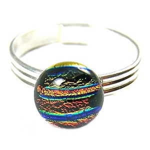 May include: A silver-toned ring with a round, dichroic glass stone. The stone features a dark base with layers of orange, blue, and green, creating a colorful, iridescent effect. The ring band has a triple-band design.