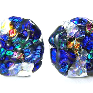 May include: Two dichroic glass beads with a unique, organic shape. The beads feature a dark base with vibrant blue, green, and iridescent accents. The glass has a textured appearance, with a mix of colors and light reflections.