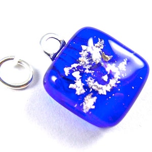 May include: A square, cobalt blue glass pendant with silver flecks and a silver bail. A separate silver jump ring is next to the pendant. The pendant is likely for a necklace or bracelet.