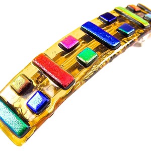 May include: A handcrafted, gold-toned glass barrette with a curved shape. It features a mosaic of colorful, rectangular and square glass pieces in shades of red, blue, green, and pink. The barrette is designed to hold hair in place.