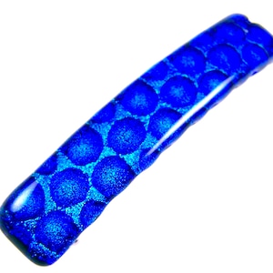May include: A rectangular, dichroic glass pendant in shades of blue. The pendant features a textured surface with a pattern of raised, circular dots. The glass has a glossy finish and is set against a white background.