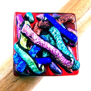 May include: A square dichroic glass ring with a red base. The ring features colorful, textured glass elements in shades of blue, purple, green, and brown. The ring is displayed on a wooden dowel.