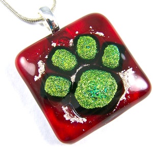 May include: A square, red glass pendant with a paw print design. The paw print is made of green, textured glass and is set against a black background. The pendant hangs from a silver chain.