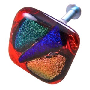 May include: A square cabinet knob made of dichroic glass, with a red border and a colourful interior. The interior has green, blue, and gold iridescent glass. A silver-coloured metal stem extends from the back.