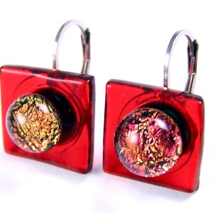 May include: A pair of red square earrings with a round, multi-coloured glass centre. The earrings have silver-coloured lever-back closures. The glass centre features gold, pink, and black flecks.