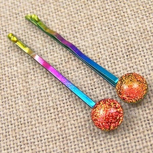May include: Two rainbow-colored bobby pins with a round, textured, iridescent bead at the end. The pins transition from yellow to blue, with a red and gold bead. Hair accessories.