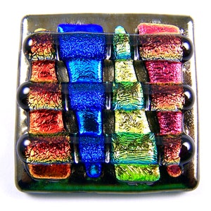 May include: Square dichroic glass pendant with a woven pattern in shades of blue, red, green, and gold. The pendant has a dark, metallic border and three black, rounded accents on each side.