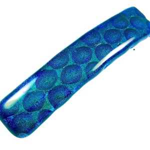May include: A rectangular, iridescent blue hair barrette with a pattern of large, circular dots. The barrette is made of glass and has a glossy finish. The barrette is approximately 3 inches long.