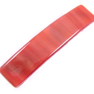 May include: A rectangular, reddish-orange hair barrette with a glossy finish. The barrette has a smooth, polished surface and a slightly curved shape, suitable for various hairstyles.
