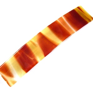 May include: A rectangular hair barrette with a glossy finish. The barrette features a striped pattern of alternating bands of amber and cream colors. The accessory is angled diagonally against a white background.