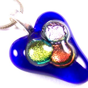 May include: A heart-shaped pendant with a vibrant blue base and three colorful, iridescent glass accents. The pendant is suspended from a silver chain, creating a striking contrast. The glass accents feature shades of green, orange, and silver.