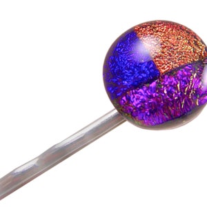 May include: A colorful hair pin with a round, dichroic glass bead. The bead is divided into three sections of blue, red-orange, and purple. The pin is silver-toned and angled to the left.