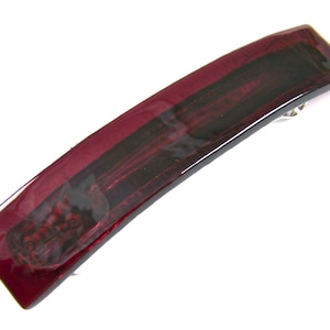 May include: A rectangular, curved hair barrette in shades of burgundy and black. The barrette has a glossy finish and a silver-colored clasp on the back. The design features a dark, textured pattern.