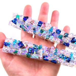 May include: Two rectangular, clear resin hair barrettes. Each barrette is embedded with iridescent, blue, green, and clear glass pieces. The barrettes are held in a hand against a white background.