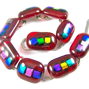 Puede incluir: Una pulsera de vidrio dicroico rojo con cuentas rectangulares. Cada cuenta presenta un cuadrado de vidrio iridiscente colorido. Las cuentas están conectadas por anillos de metal plateado, creando un accesorio único.