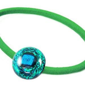 May include: A green hair tie with a round, dichroic glass bead. The bead features a mix of blue and green colors, creating a shimmering effect. The hair accessory is designed for styling hair.