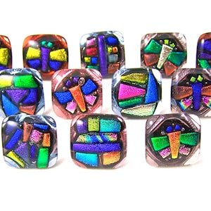 May include: A collection of square glass beads with dichroic glass designs. Each bead features a unique mosaic of colors including purple, green, yellow, and orange. The beads are arranged on a white surface, showcasing their vibrant, reflective qualities.