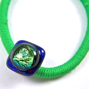 May include: A green hair tie with a square, dichroic glass bead. The bead has a dark blue border and a center with green and gold iridescent colors. The hair tie is made of a thick, textured green fabric.