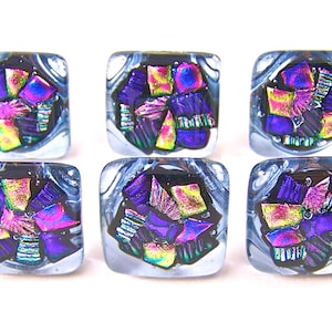May include: Six square glass beads with dichroic glass inserts. The beads feature a mix of purple, pink, yellow, and green colors. The glass has a swirling, marbled effect around the edges.