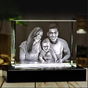 Peut inclure: Un bloc de cristal transparent et rectangulaire avec un portrait de famille gravé à l'intérieur. L'image montre un couple souriant et un bébé. Le cristal repose sur une base noire éclairée, créant un effet lumineux.
