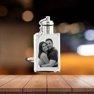 以下が含まれることがあります： シルバーカラーのトップと金属製のループが付いた、透明な長方形のキーホルダー。キーホルダーには、笑顔のカップルが抱き合っている白黒写真が入っています。背景はぼやけています。