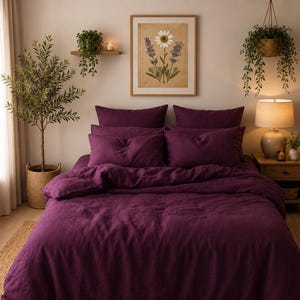 Op de afbeelding: Een dieppaarse beddengoedset bestaande uit een dekbedovertrek en vier bijpassende kussens. De stof heeft een getextureerd, mat uiterlijk. Het bed staat centraal in een kamer met een ingelijste botanische print van een madeliefje en lavendel.