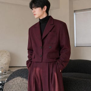Puede incluir: Una chaqueta corta burdeos con dos bolsillos delanteros y pantalones de cintura alta a juego. La chaqueta tiene solapas con muescas y dos botones. El conjunto se combina con un cuello alto negro. El conjunto es una versión moderna de un traje clásico.