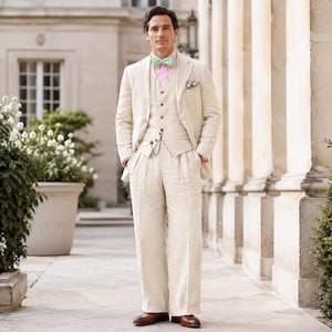 May include: A three-piece beige linen suit with a matching vest and wide-leg trousers. The suit is paired with a pink dress shirt, a light green bow tie, and brown leather shoes. A pocket square adds a touch of detail to the ensemble.