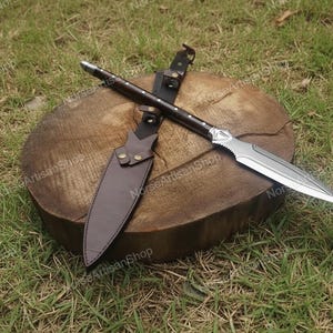 Puede incluir: Una lanza artesanal con una hoja de acero pulido y un mango de madera oscura, apoyada sobre una superficie de madera. Tambi&eacute;n se ven una funda y correas de cuero marr&oacute;n. La lanza est&aacute; dise&ntilde;ada para la recreaci&oacute;n hist&oacute;rica o la exhibici&oacute;n.