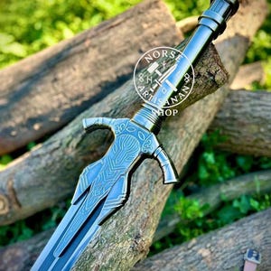 May include: A detailed, blue-grey sword with intricate designs on the hilt and blade, resting on wooden logs. The sword features a crossguard and a pointed tip. The image includes the text "NORS ARTISAN SHOP" in a circular logo.