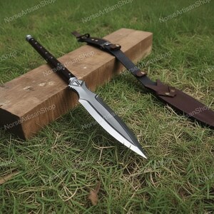 Puede incluir: Una lanza plateada y negra con funda y mango de cuero descansa sobre un bloque de madera y hierba. La lanza tiene una hoja afilada y puntiaguda y un mango decorativo. La funda es marr&oacute;n y tiene una correa.