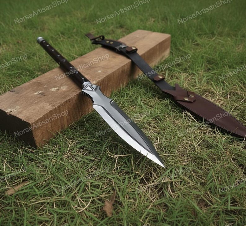 Puede incluir: Una lanza plateada y negra con funda y mango de cuero descansa sobre un bloque de madera y hierba. La lanza tiene una hoja afilada y puntiaguda y un mango decorativo. La funda es marr&oacute;n y tiene una correa.