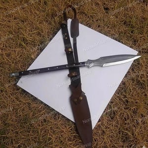 Puede incluir: Una lanza con un asta de madera oscura y una hoja plateada, sobre una superficie blanca. La lanza va acompa&ntilde;ada de una funda de cuero y una correa de transporte. El dise&ntilde;o sugiere un tema hist&oacute;rico o de fantas&iacute;a.