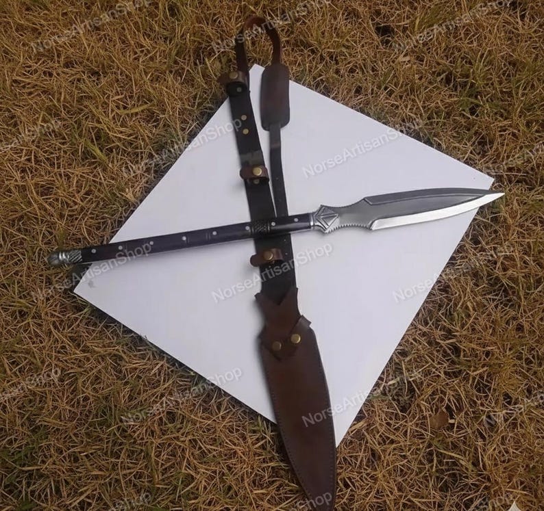 Puede incluir: Una lanza con un asta de madera oscura y una hoja plateada, sobre una superficie blanca. La lanza va acompa&ntilde;ada de una funda de cuero y una correa de transporte. El dise&ntilde;o sugiere un tema hist&oacute;rico o de fantas&iacute;a.