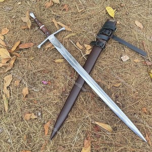Puede incluir: Una espada larga y plateada con un mango de madera marrón y una guarda cruzada, sobre un lecho de hierba seca y hojas. La espada se combina con una funda de cuero marrón con correas negras.