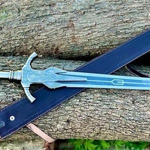 May include: A silver sword with a detailed hilt and a blue blade rests on a blue sheath. The sword is placed on a wooden surface. The image includes the text "Norse Artisans Shop" in a circular logo.