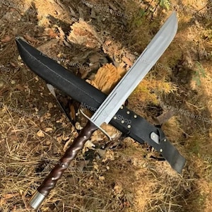 May include: A large, silver-bladed sword with a brown, textured handle and a black sheath. The sword has a crossguard and a polished metal finish. The sheath is made of black leather. The sword is lying on a bed of brown leaves and twigs.