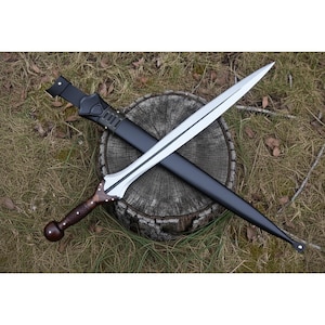 Puede incluir: Una espada plateada con una hoja pulida y un mango de madera oscura con un pomo redondeado. La espada descansa en una vaina de cuero negro, con una trabilla de cinturón de cuero negro. La espada y la vaina están colocadas sobre un tocón.