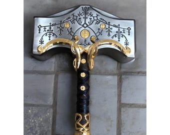 May include: A detailed, metallic hammer with intricate gold and silver detailing. The hammerhead features engraved patterns and circular accents. The handle is wrapped in black leather with gold accents at the base. A decorative piece.