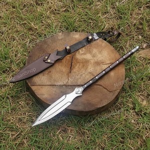 Puede incluir: Una lanza y un cuchillo con funda de cuero descansan sobre una superficie de madera. La lanza tiene una hoja plateada y un mango marr&oacute;n oscuro con detalles plateados. El cuchillo tiene una funda de cuero marr&oacute;n y un mango negro.