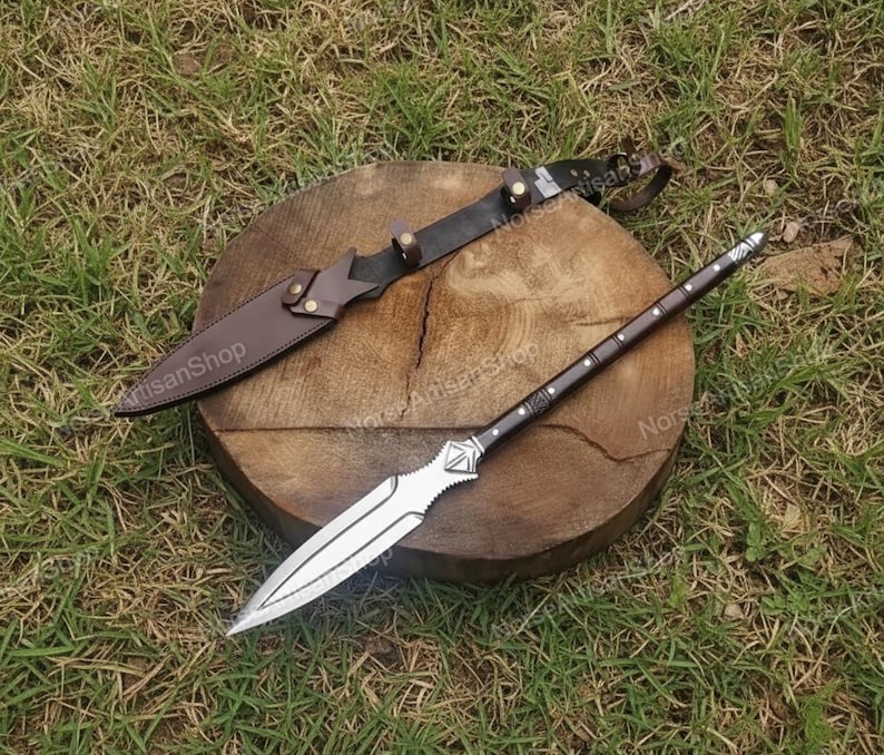 Puede incluir: Una lanza y un cuchillo con funda de cuero descansan sobre una superficie de madera. La lanza tiene una hoja plateada y un mango marr&oacute;n oscuro con detalles plateados. El cuchillo tiene una funda de cuero marr&oacute;n y un mango negro.