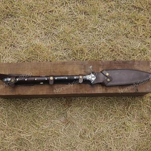 Puede incluir: Una lanza artesanal con una funda y un mango de cuero marr&oacute;n oscuro. La punta de la lanza es plateada con un dise&ntilde;o geom&eacute;trico. El mango est&aacute; envuelto en cuero marr&oacute;n oscuro con detalles plateados. La lanza est&aacute; sobre un bloque de madera.