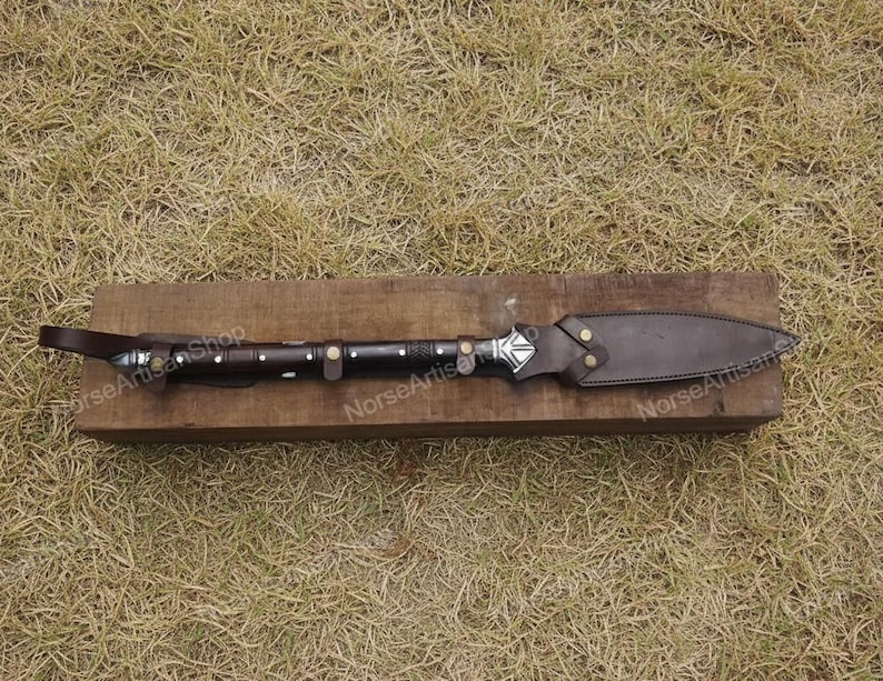 Puede incluir: Una lanza artesanal con una funda y un mango de cuero marr&oacute;n oscuro. La punta de la lanza es plateada con un dise&ntilde;o geom&eacute;trico. El mango est&aacute; envuelto en cuero marr&oacute;n oscuro con detalles plateados. La lanza est&aacute; sobre un bloque de madera.