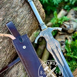 May include: A detailed image of a sword with a silver blade and handle, resting against a tree trunk. The sword has intricate designs on the hilt and a leather sheath. The image also includes the text "NORSE ARTISAN SHOP" in a circular logo.