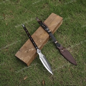 Puede incluir: Una lanza con una hoja plateada y un mango de madera oscura, apoyada sobre un bloque de madera. Tambi&eacute;n hay una funda y una correa de cuero. La lanza est&aacute; dise&ntilde;ada con una punta afilada y un mango decorativo.