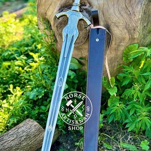 May include: A silver and blue sword with intricate designs, resting beside a dark blue leather sheath. The sword features a detailed hilt and blade, with the "Norse Artisan Shop" logo visible. The scene is set outdoors with green foliage.
