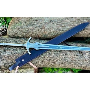 May include: A silver-colored sword with a decorative hilt and a long, light blue blade rests on a dark blue sheath. The sword is displayed on a bed of wooden logs. The logo for Norse Artisan Shop is visible in the upper left corner.